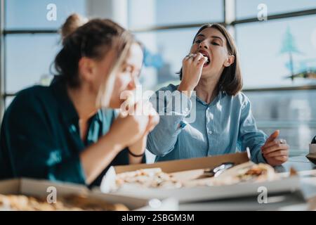 Two cheerful colleagues taking a lunch break in the office kitchen, enjoying fresh pizza and relaxing together. Stock Photo