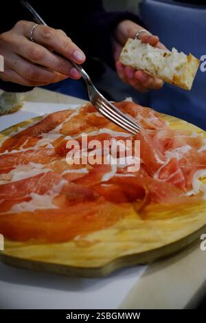 Thinly sliced cured meat arranged on a wooden board, alongside a piece of crusty bread. A person uses a fork to prepare a bite. Stock Photo