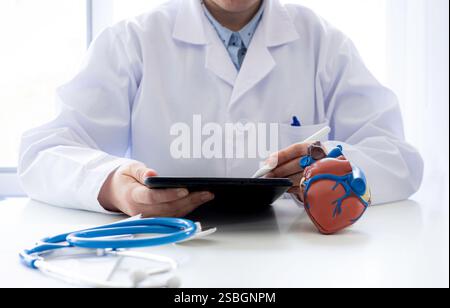 Doctor in a gown using a digital tablet with a stylus. On the table is a model of a heart and a stethoscope, representing modern cardiology, digital Stock Photo