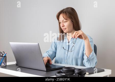A calm nice woman with eyeglasses in her hand is typing on the keyboard using laptop, looking at PC screen, remote hardworking freelancer works from h Stock Photo