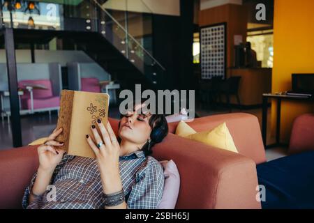 A young female college student enjoys reading a book while relaxing on a comfortable couch in a modern lounge area. Stock Photo