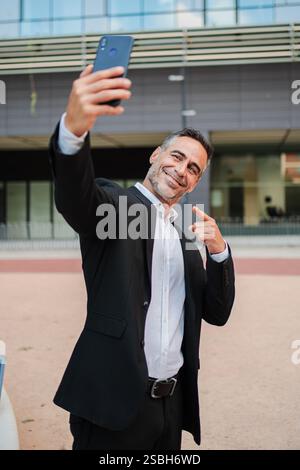 A Confident Businessman Takes A Selfie With His Smartphone, Smiling Brightly And Pointing At The Camera. This Engaging Pose Captures A Moment Of Joy, Success, And Communication In A Business Context. High quality photo Stock Photo