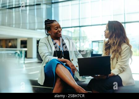 Two professional female colleagues engage in conversation while seated in a bright, modern lobby. They are sharing ideas and working on a laptop, refl Stock Photo