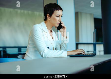 A professional woman deeply engaged in her work, using a laptop and smartphone in a modern office. Her focused expression and stylish white blazer con Stock Photo