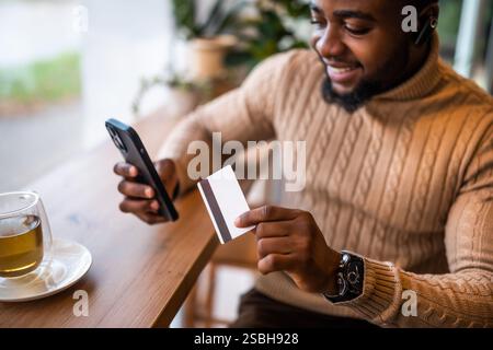 Young man in casual clothing using credit card and smartphone while sitting in the bar and drinking tea. Stock Photo
