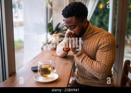 Young man sitting in bar, relaxing and drinking tea. He is blowing nose. Stock Photo