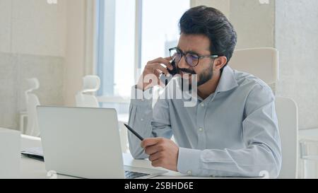 Busy multitasking Arabian businessman manager consultant Indian muslim man talk mobile phone check information on laptop computer work consulting Stock Photo