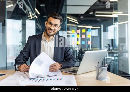 Portrait of successful financier, man signs documents reports contracts. Businessman at workplace inside office doing paper work, filling out tax and statistical forms, looking at camera smiling. Stock Photo