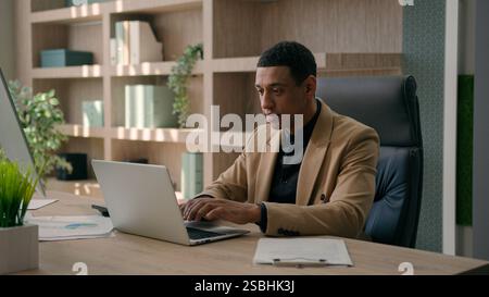 Smiling businessman African American man male entrepreneur business leader software engineer typing project online on laptop in office workplace happy Stock Photo