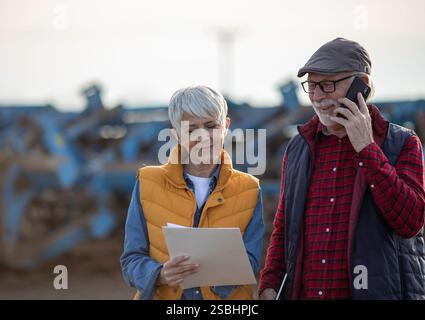 Mature farmers man and woman standing in field, writing notes and talking on mobile phone while tractor plowing in background Stock Photo