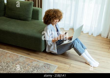 Seated comfortably on the floor, a young woman engages with her laptop while checking her smartphone. The cozy, sunlit living room creates a serene ba Stock Photo