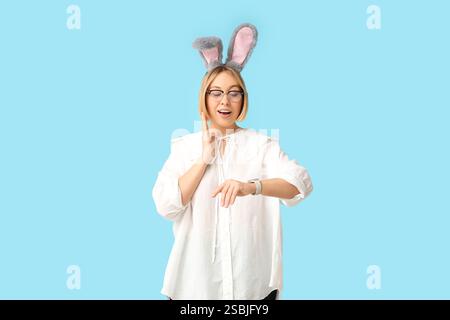 Worried adult businesswoman in bunny ears looking at wristwatch on blue background. Easter celebration Stock Photo