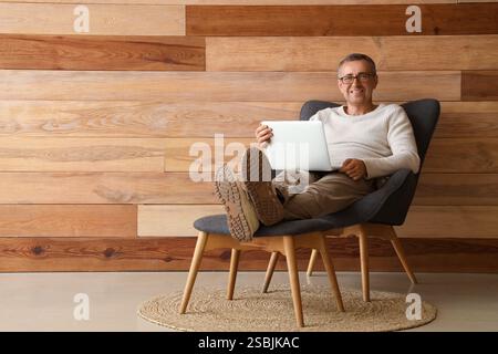 Old man sitting in armchair with legs on footstool and using laptop near wooden wall Stock Photo