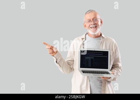 Mature programmer with laptop pointing at something on light background Stock Photo
