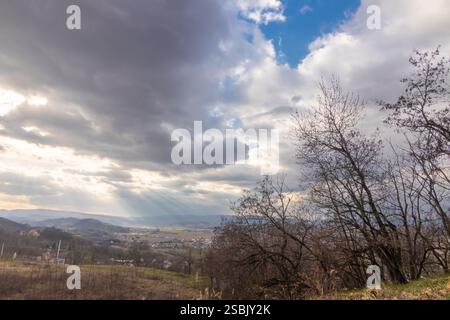 Dramatic rural scenery featuring rolling hills, dramatic clouds breaking into sun rays, and silhouetted trees. Captured under dynamic lighting conditi Stock Photo