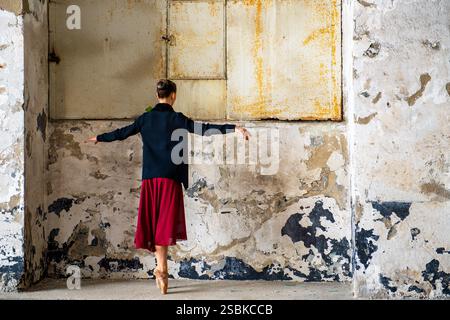 A ballet dancer in a red skirt and black sweater gracefully balancing en pointe in an abandoned industrial space, creating a contrast between elegance Stock Photo