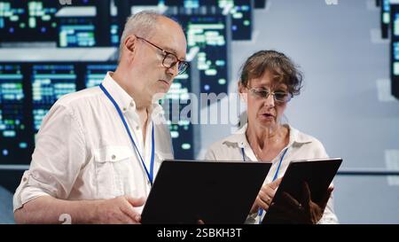 Data storage company executives looking around data center, using laptop and tablet to crosscheck contingency plan and assess high tech workspace units in need of fixing, preventing liabilities Stock Photo