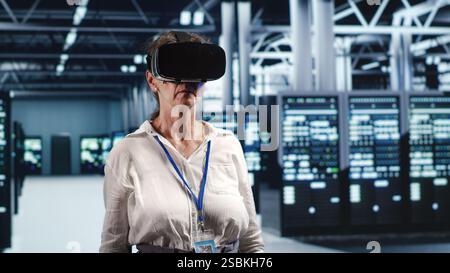 Older computer scientist in modern server hub wearing virtual reality headset, performing racks maintenance. Licensed professional using vr glasses in high tech workplace to do checkup on hardware Stock Photo