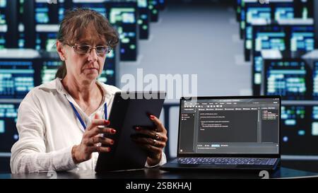 Elderly woman working in IT in high tech workplace completing tasks on tablet while running scripts on laptop terminal. Professional amidst server cabinets manipulates code and improves cybersecurity Stock Photo