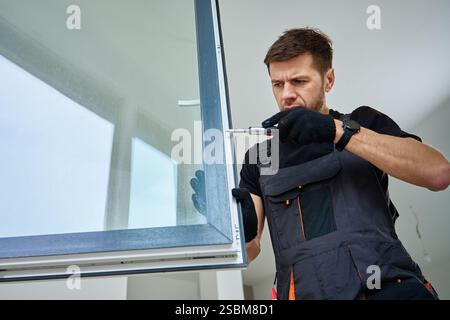 Skilled worker wearing gloves uses screwdriver to fix window frame, home renovation tasks. Professional repairman adjusting white pvc windows in moder Stock Photo