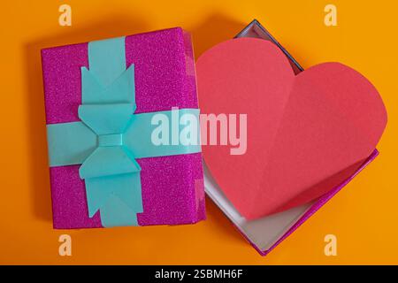 Top view of an open gift box with a red paper heart on a bold orange background, symbolizing love, gift, appreciation, and celebration for Valentine's Stock Photo