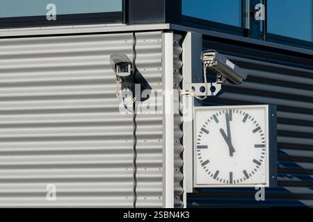 Two security cameras are positioned on a contemporary building's façade alongside a large, round clock showing the time. The structure features sleek Stock Photo