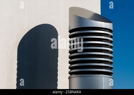 Modern ventilation cylindrical pipe stands against a clear blue sky, its spiral design creating an interesting shadow on the adjacent white wall in br Stock Photo