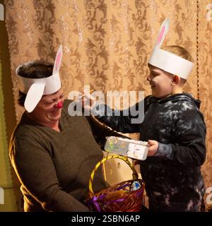 A boy and an adult woman, both wearing bunny ear headbands, participate in an Easter activity. The boy playfully paints the woman's face. Stock Photo