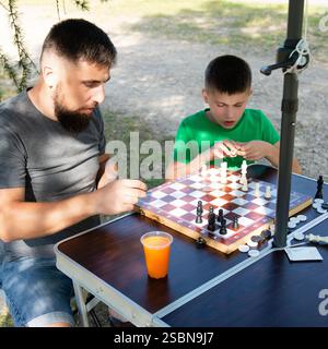 A man and a boy are playing chess outdoors on a portable table. They are both attentively focused on the game, enjoying a moment of shared activity in Stock Photo