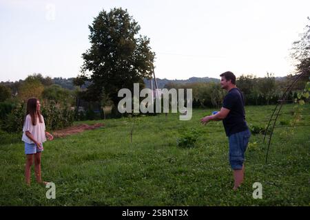 A man and a girl engage in a game of catch in a lush green yard. The setting sun casts a warm glow. Trees and a hillside form the backdrop. Stock Photo