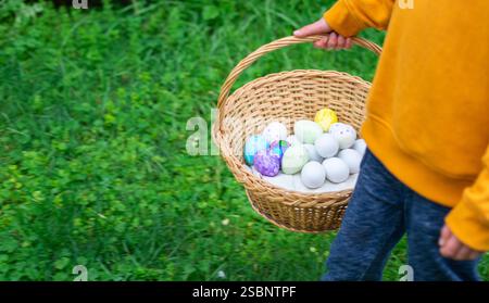 Kid holding wicker basket full of colorful decorated and white easter eggs walking in a green grass field during easter egg hunt Stock Photo