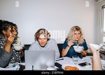 Male student using laptop while female roommates eating noodles sitting at desk in college dorm Stock Photo