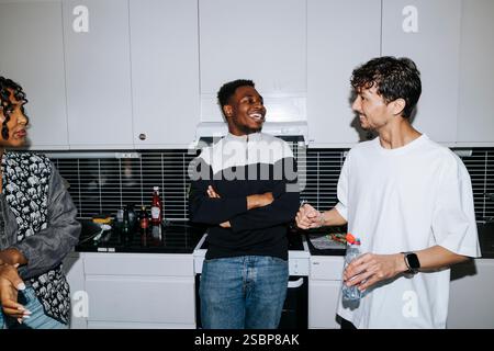 Young man holding water bottle and talking with male and female roommates standing in dorm room kitchen Stock Photo