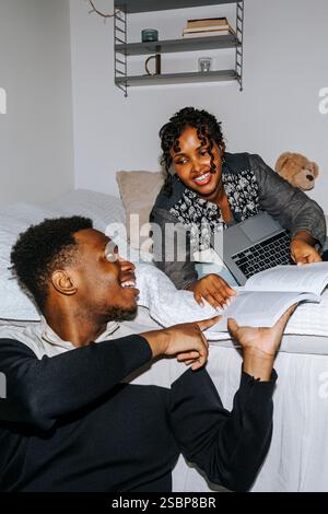 Young man showing book to female roommate lying down on bed in college dorm room Stock Photo
