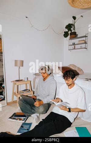 Young male roommates studying together while sitting by bed in college dorm Stock Photo