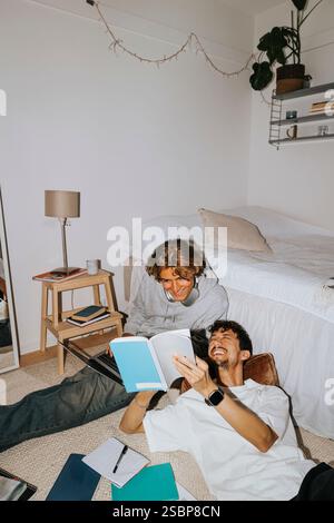 Happy young man showing book to male friend sitting by bed in college dorm Stock Photo