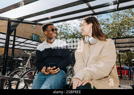 Happy multiracial male and female friends talking while sitting on railing at bicycle parking station Stock Photo