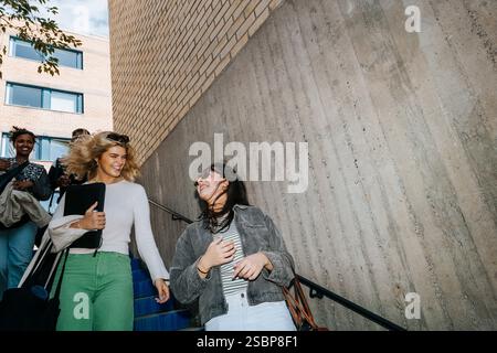 Low angle view of happy female students talking while moving down from stairs Stock Photo