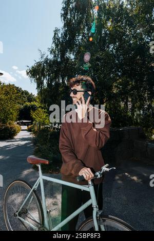 Smiling young man talking on smart phone while standing with bicycle on footpath at sunny day Stock Photo