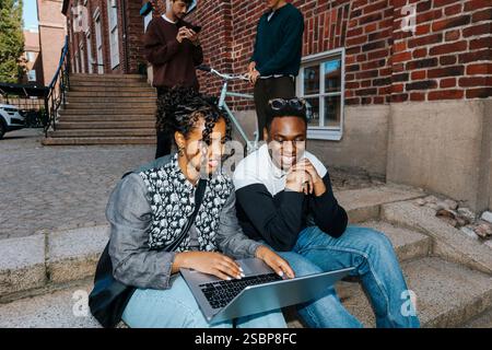 Young female student using laptop while sitting with male friend on steps at college campus Stock Photo