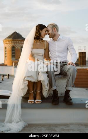 Smiling young man sitting with bride wearing veil while sitting on rooftop Stock Photo
