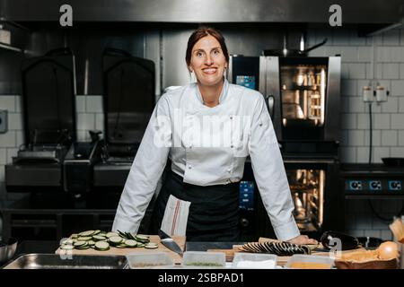 Portrait of smiling female chef wearing white uniform and standing near counter in kitchen at restaurant Stock Photo