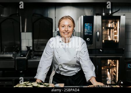 Portrait of smiling young female chef wearing white uniform and leaning on counter in kitchen at restaurant Stock Photo
