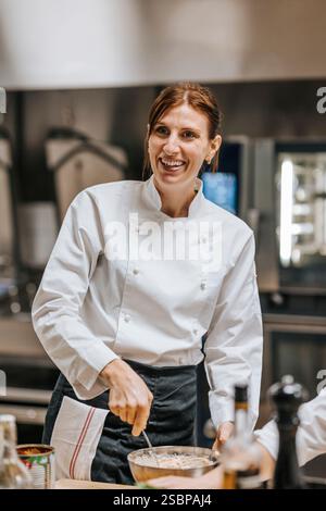 Happy female chef in white uniform mixing batter while standing in commercial kitchen Stock Photo