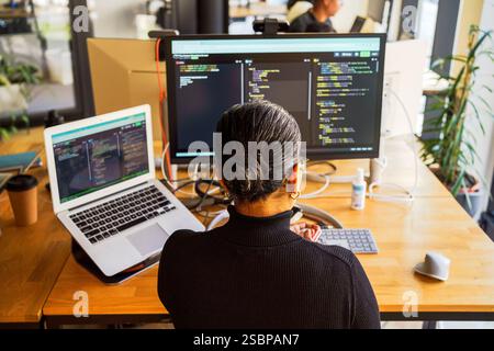 High angle rear view of female programmer coding on desktop PC and laptop while sitting at desk in office Stock Photo