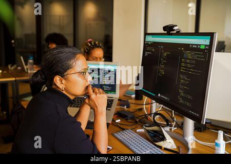 Thoughtful female coder looking at desktop PC while sitting in startup office Stock Photo
