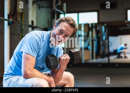 A senior man smiles as he sits on a bench and lifts a dumbbell He is working his biceps in a gym Stock Photo