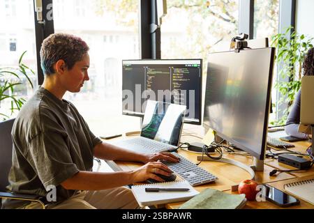 Focused non-binary IT professional working on desktop PC at desk in startup office Stock Photo