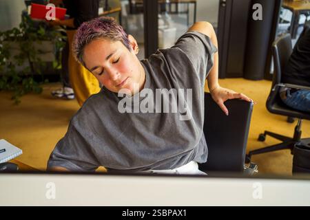 Tired non-binary programmer stretching while sitting on chair in office Stock Photo