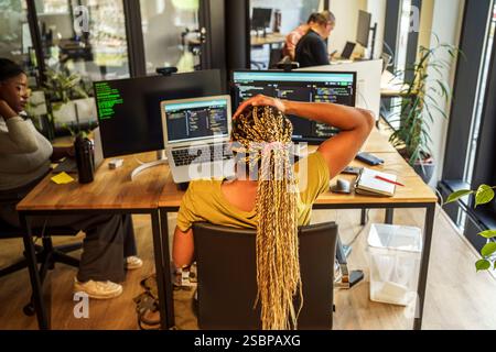 High angle rear view of braided hair female coding professional sitting near desktop PC and laptop in tech hub Stock Photo